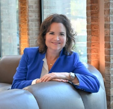 Professional woman in blue blazer smiling at camera while seated indoors with brick wall background